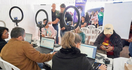 A imagem mostra um grupo de pessoas trabalhando em mesas brancas com notebooks em um espaço cobe...