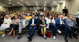 Fotografia em plano aberto mostra um auditório cheio, com dezenas de pessoas sentadas em cadeira...