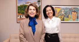 Fotografia de duas mulheres sorridentes em pé, lado a lado. A mulher à esquerda tem cabelos curt...
