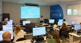 Fotografia colorida de uma sala de informática clara e moderna. Várias pessoas adultas estão sen...
