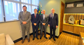Quatro homens de terno estão em pé lado a lado em um escritório institucional, posando para uma ...