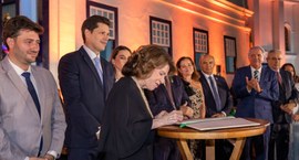Uma mulher de cabelo castanho curto e terno preto assina um livro de atas sobre uma mesa redonda...