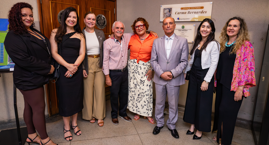 Oito pessoas com roupas formais e casuais posam sorridentes em um ambiente interno, posicionadas...