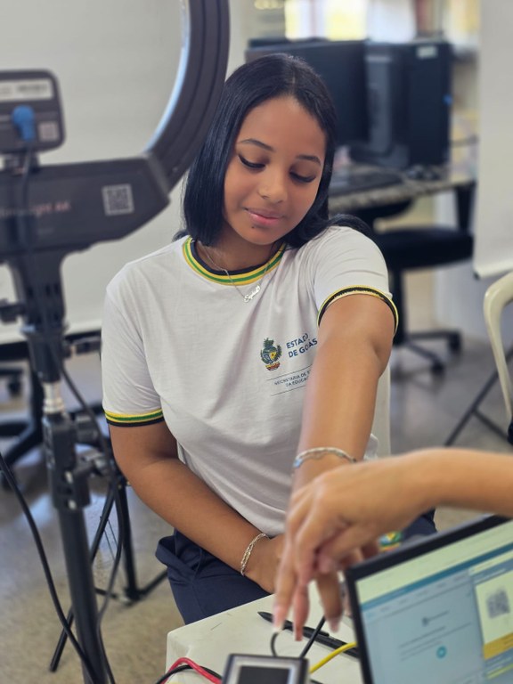 Uma jovem estudante, com camiseta branca do Estado de Goiás, está sentada e estende o braço sobr...