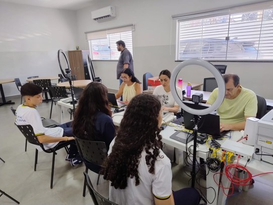 Sala clara com várias mesas e computadores. Alguns estudantes estão sentados enquanto servidores...