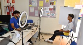 A imagem mostra um atendimento sendo realizado em um ambiente interno. Um homem está sentado atr...