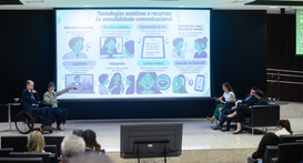 A imagem retrata um auditório durante uma palestra ou evento voltado à inclusão, com a plateia e...