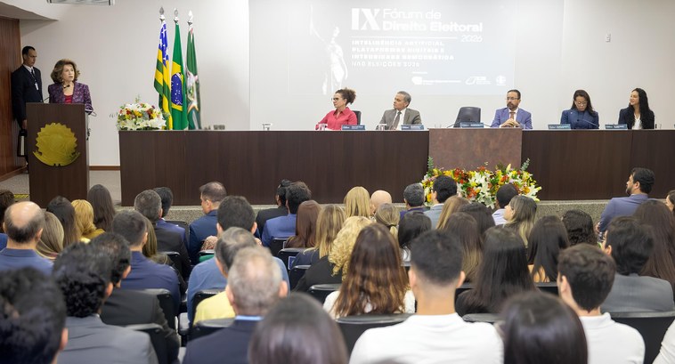 Auditório cheio durante um evento institucional: à frente da sala, uma pessoa fala em um púlpito...