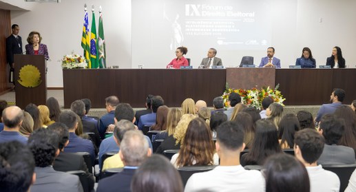 Auditório cheio durante um evento institucional: à frente da sala, uma pessoa fala em um púlpito...