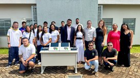 A imagem mostra um grupo de cerca de 20 pessoas reunidas ao ar livre, em frente a um prédio de p...