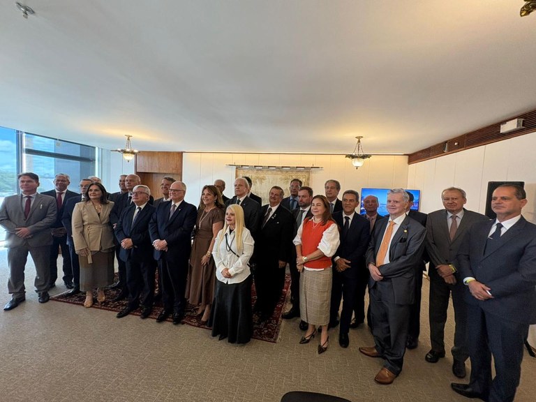 Uma foto em grupo composta pessoas em pé, posicionadas em um ambiente amplo e formal. A maioria ...