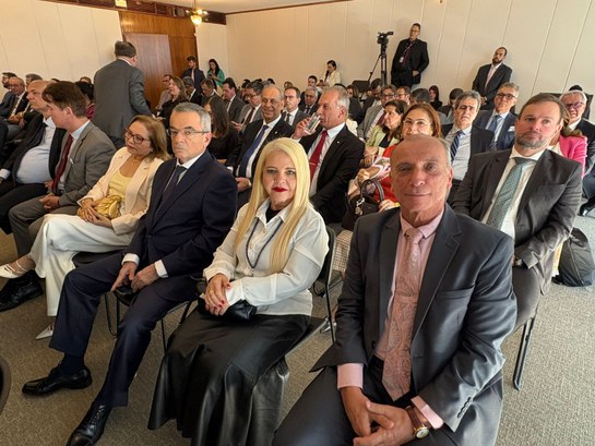 A foto mostra um auditório ou sala de reuniões com várias pessoas sentadas em fileiras, vestindo...