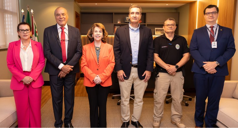Fotografia colorida de seis pessoas posando de pé, lado a lado, em um escritório moderno com dec...