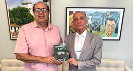 A imagem apresenta dois homens em pé, do peito para cima, posicionados em um ambiente interno de...