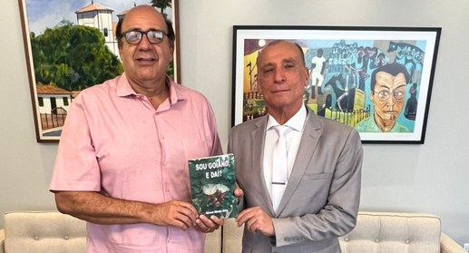 A imagem apresenta dois homens em pé, do peito para cima, posicionados em um ambiente interno de...