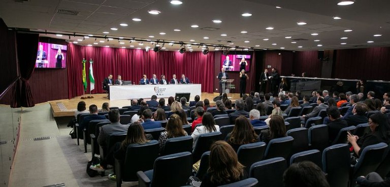 Fotografia do palco de um auditório em que um homem aparece falando em um púlpito, acompanhado d...