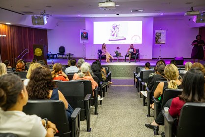 Fotografia de duas mulheres sentadas em poltronas pretas num palco durante um evento. Em primeir...