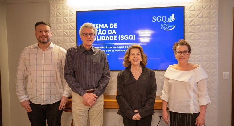 Uma fotografia em ambiente interno mostra quatro pessoas em pé, lado a lado e sorrindo para a câ...