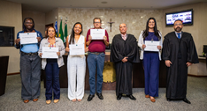 Fotografia de sete pessoas posicionadas lado a lado no plenário de um tribunal. Quatro mulheres ...