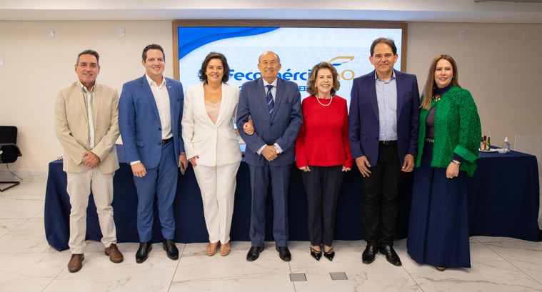 A imagem mostra um grupo de sete pessoas, cinco homens e duas mulheres, de pé em linha e sorrind...