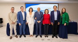 A imagem mostra um grupo de sete pessoas, cinco homens e duas mulheres, de pé em linha e sorrind...