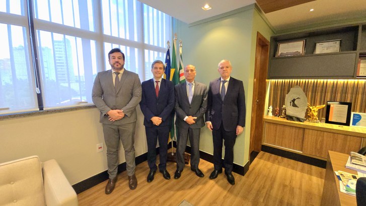 Quatro homens de terno estão em pé lado a lado em um escritório institucional, posando para uma ...