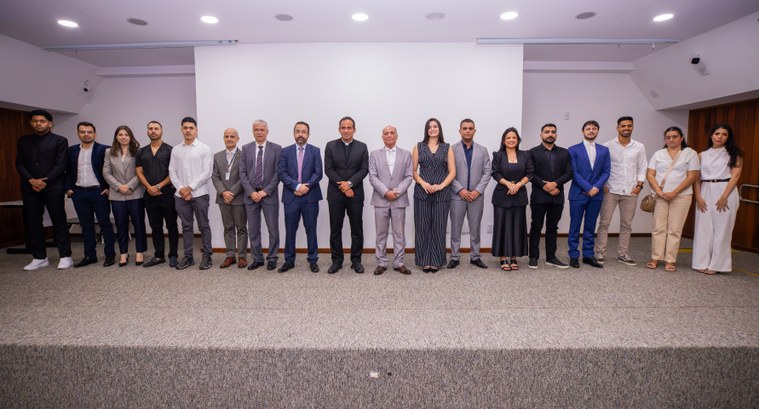 Uma fotografia em plano médio e horizontal captura um grupo de 18 pessoas perfiladas lado a lado...