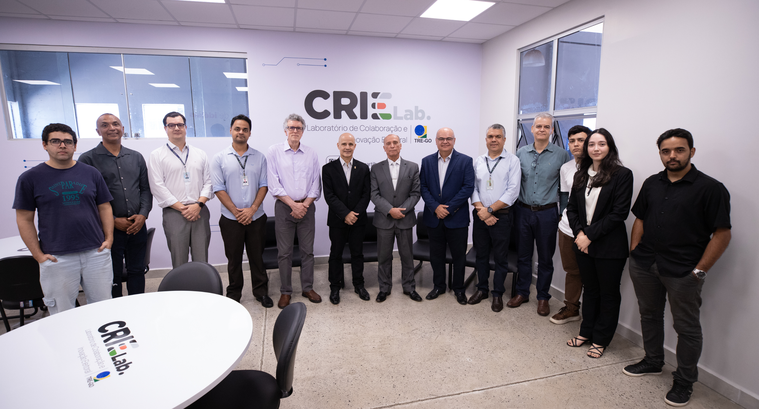 Foto em ambiente interno mostrando um grupo de treze pessoas em pé, alinhadas lado a lado, posan...