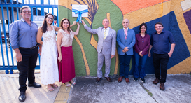 Uma fotografia em plano médio mostra sete pessoas em pé, lado a lado, em frente a um muro colori...