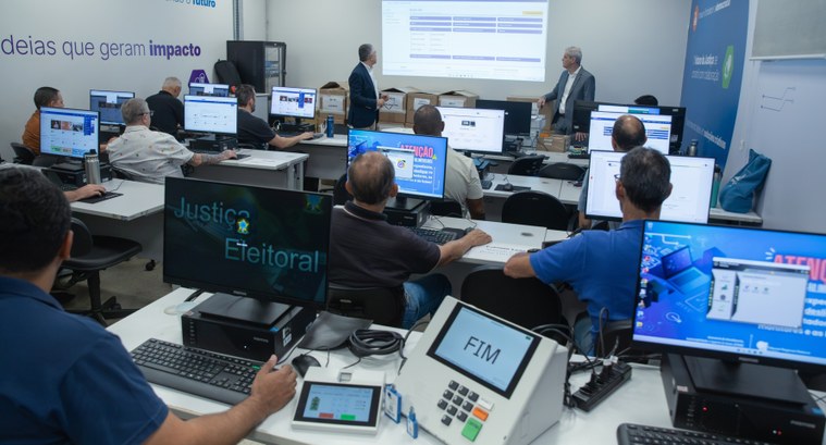 Pessoas sentadas em uma sala de informática utilizam computadores e urnas eletrônicas durante um...