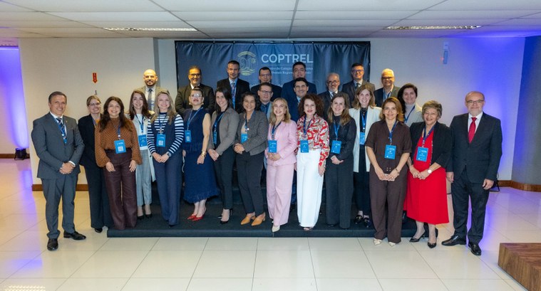 Grupo de aproximadamente 25 pessoas posa para uma foto em um ambiente interno de evento corporat...