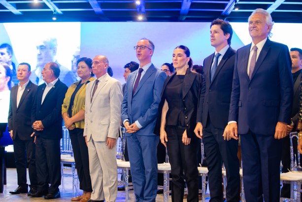 Em um palco montado para a solenidade, autoridades estão alinhadas em pé, compondo o dispositivo...