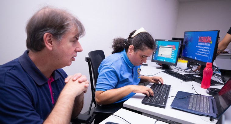 Um homem e uma mulher estão sentados lado a lado, de frente para computadores em um ambiente de ...
