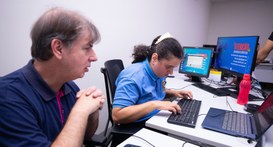Um homem e uma mulher estão sentados lado a lado, de frente para computadores em um ambiente de ...