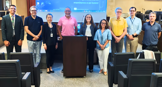 Uma fotografia em plano médio, capturada de frente, mostra nove pessoas, um painel e cadeiras em...
