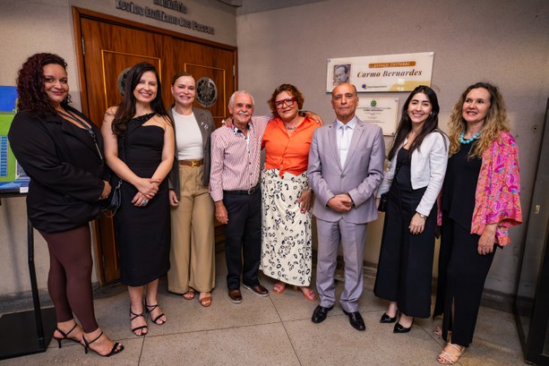 Oito pessoas com roupas formais e casuais posam sorridentes em um ambiente interno, posicionadas...