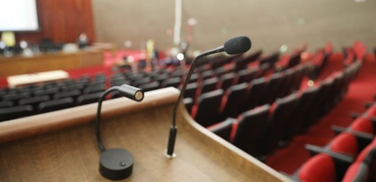TSE realizará série de audiências públicas para aperfeiçoar resoluções do processo eleitoral Imagem focada em um microfone de mesa preto e ao fundo um auditório com poltronas vermelhas