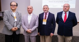 Uma fotografia interna mostra quatro homens em pé, lado a lado, em frente a uma tela de projeção...