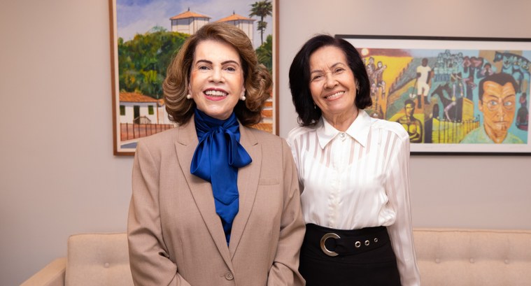 Fotografia de duas mulheres sorridentes em pé, lado a lado. A mulher à esquerda tem cabelos curt...
